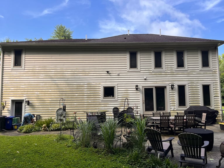 Two-story beige house with dark roof and patio area featuring black Adirondack chairs and garden plants