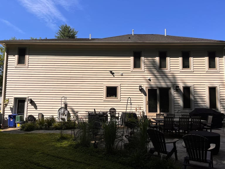 Two-story beige house with white trim, black chairs and table on patio, lawn and landscaping in foreground, clear blue sky