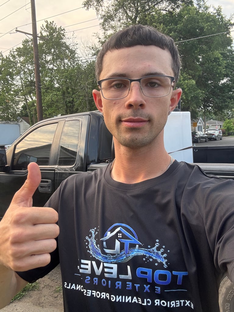 Man wearing glasses and dark t-shirt giving thumbs up gesture outdoors with parked vehicles in background