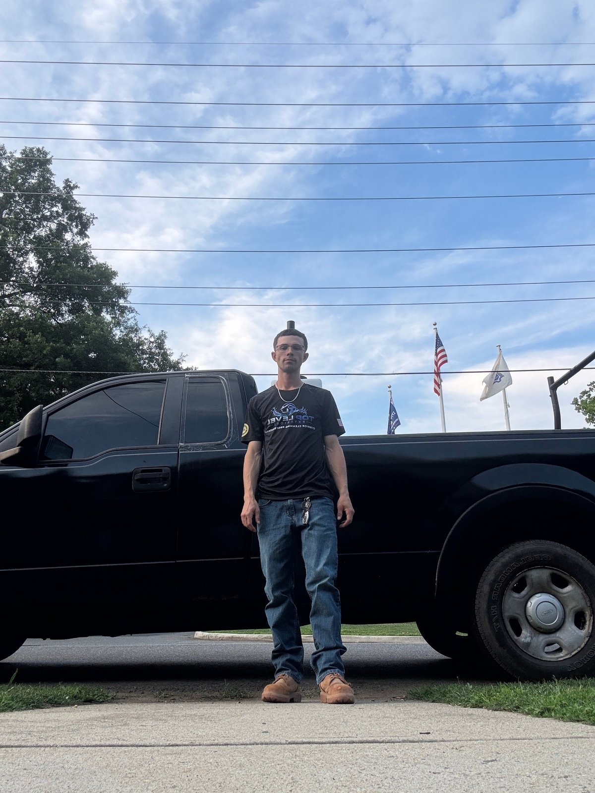 Man in dark shirt and jeans standing in front of black pickup truck with flags in background