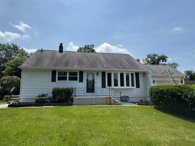 White single-story residential house with front porch, black shutters, and manicured lawn under blue sky